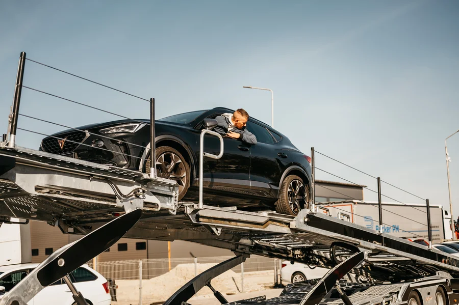 Ciemny samochód na podniesionej platformie autotransportera z osobą wychylającą się przez boczne okno.