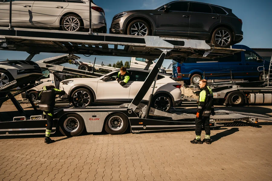 Trzy osoby przy białym samochodzie na autotransporterze; jedna znajduje się w aucie, a dwie stoją przy zestawie.