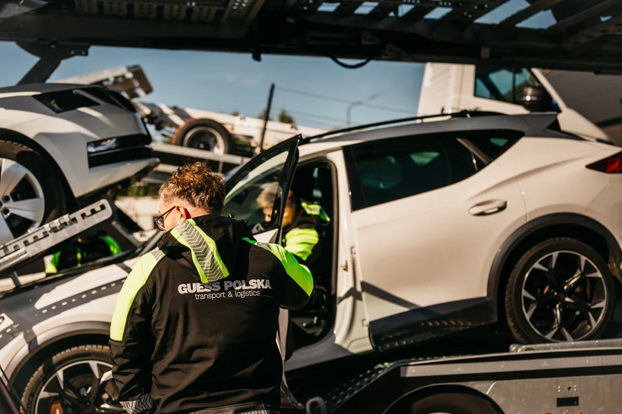 Dwie osoby przy białym samochodzie na dolnej platformie załadowanego autotransportera.
