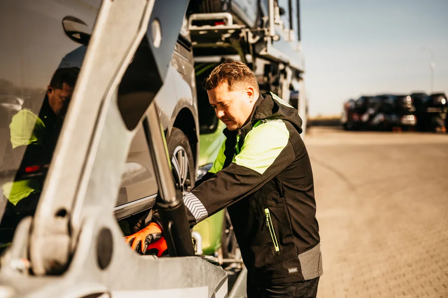 Zbliżenie na osobę w odzieży roboczej i pomarańczowych rękawicach pracującą obok samochodu i konstrukcji autotransportera.