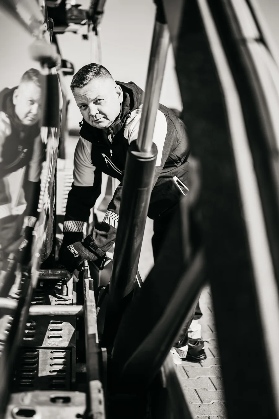 Czarno-biały pionowy portret osoby w odzieży roboczej pomiędzy samochodem a konstrukcją autotransportera.