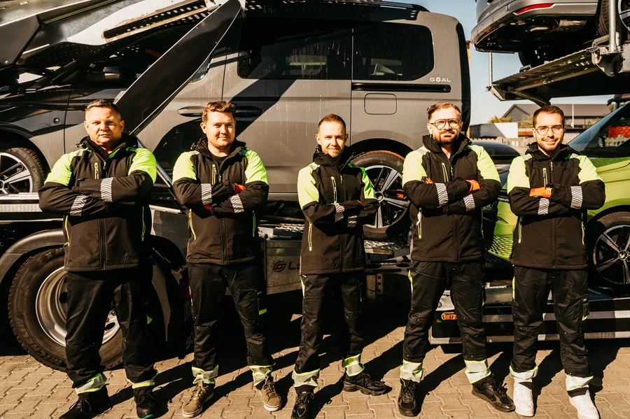Pięć osób z zespołu Guess Polska w odzieży roboczej przed autotransporterem z samochodami.