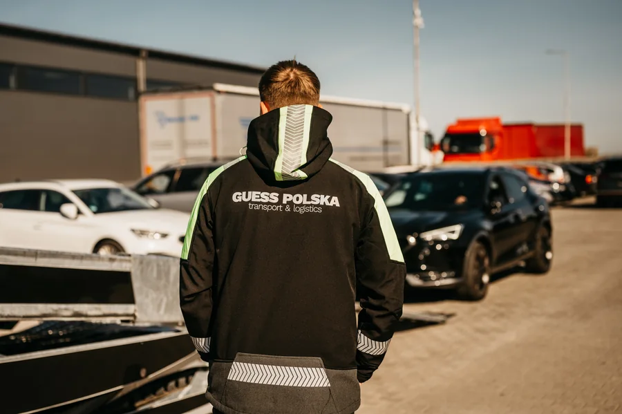 Osoba widziana od tyłu w kurtce z napisem Guess Polska, na tle samochodów i zabudowań placu.