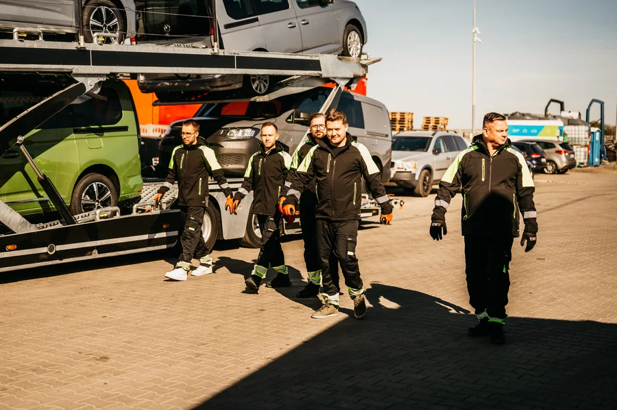 Grupa osób w odzieży Guess Polska idąca po placu obok załadowanego autotransportera.