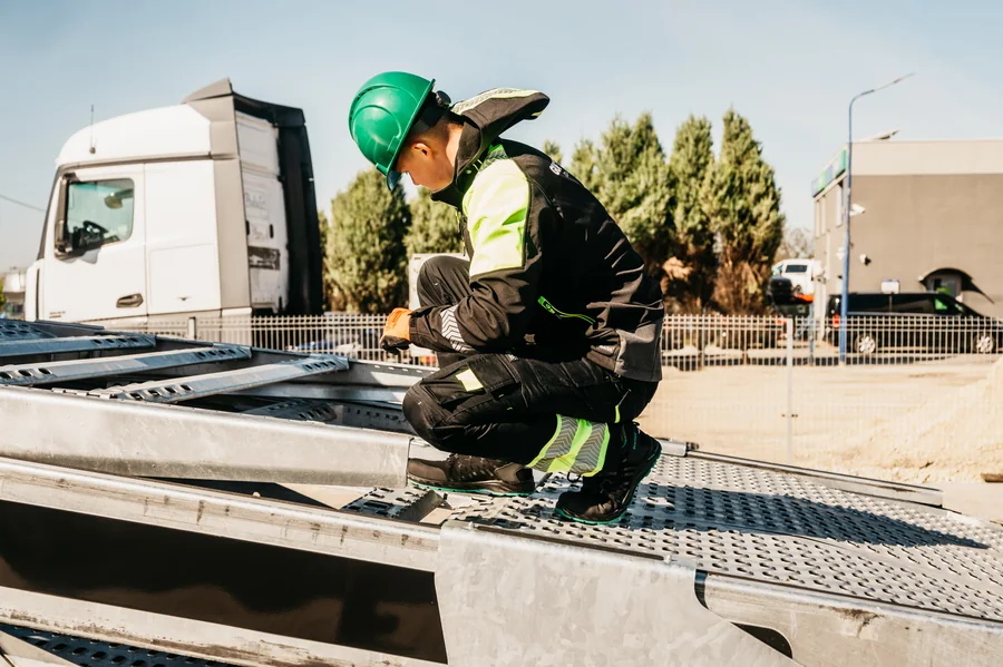 Osoba w zielonym kasku i rękawicach podczas pracy na metalowej platformie autotransportera.
