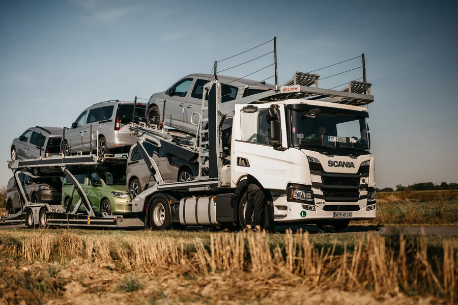 Biały autotransporter Guess Polska z samochodami na dwóch poziomach, sfotografowany z boku na tle nieba i pól.